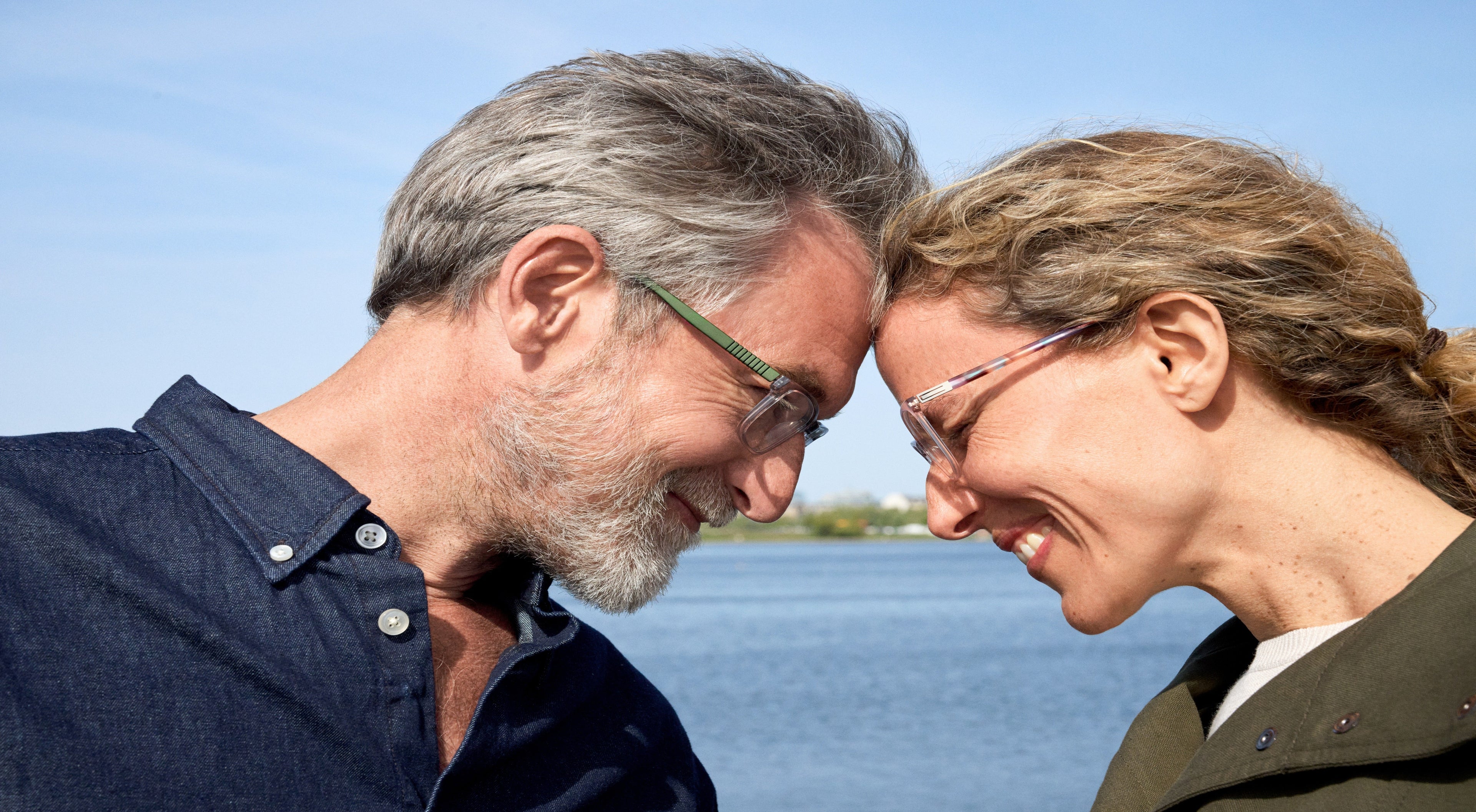 A smiling older couple gently touch foreheads while standing by the water on a sunny day, both wearing glasses and looking happy together.