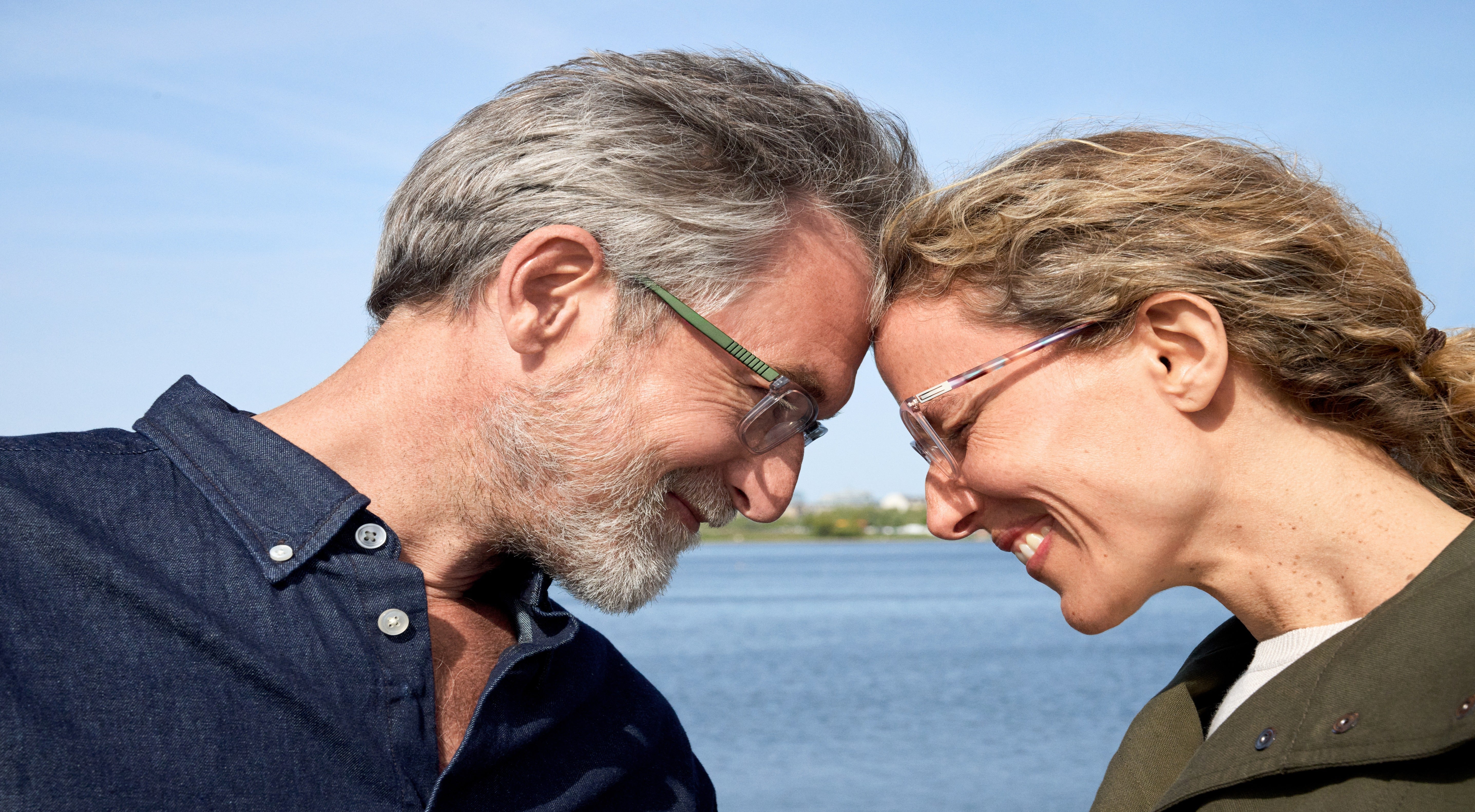 A smiling older couple gently touch foreheads while standing by the water on a sunny day, both wearing glasses and looking happy together.