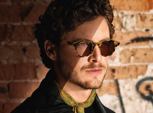 A man with curly hair and a beard wears brown tortoiseshell sunglasses with 1.5 XTRactive lenses and a green patterned scarf, standing against a graffiti brick wall as sunlight casts shadows, offering προστασία από το έντονο φως.